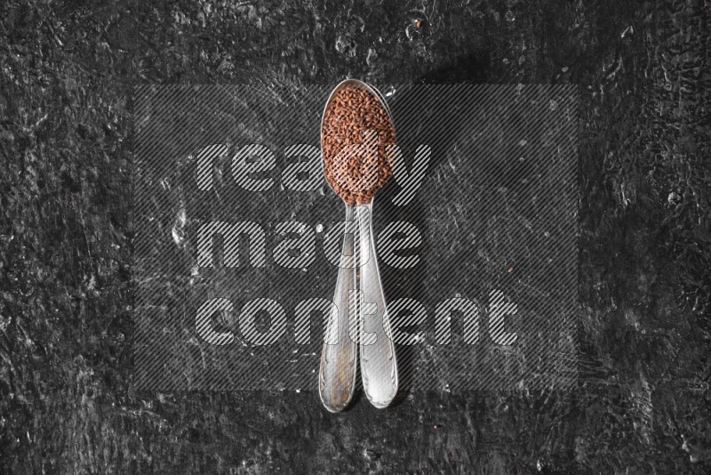2 metal spoons full of garden cress seeds on a textured black flooring