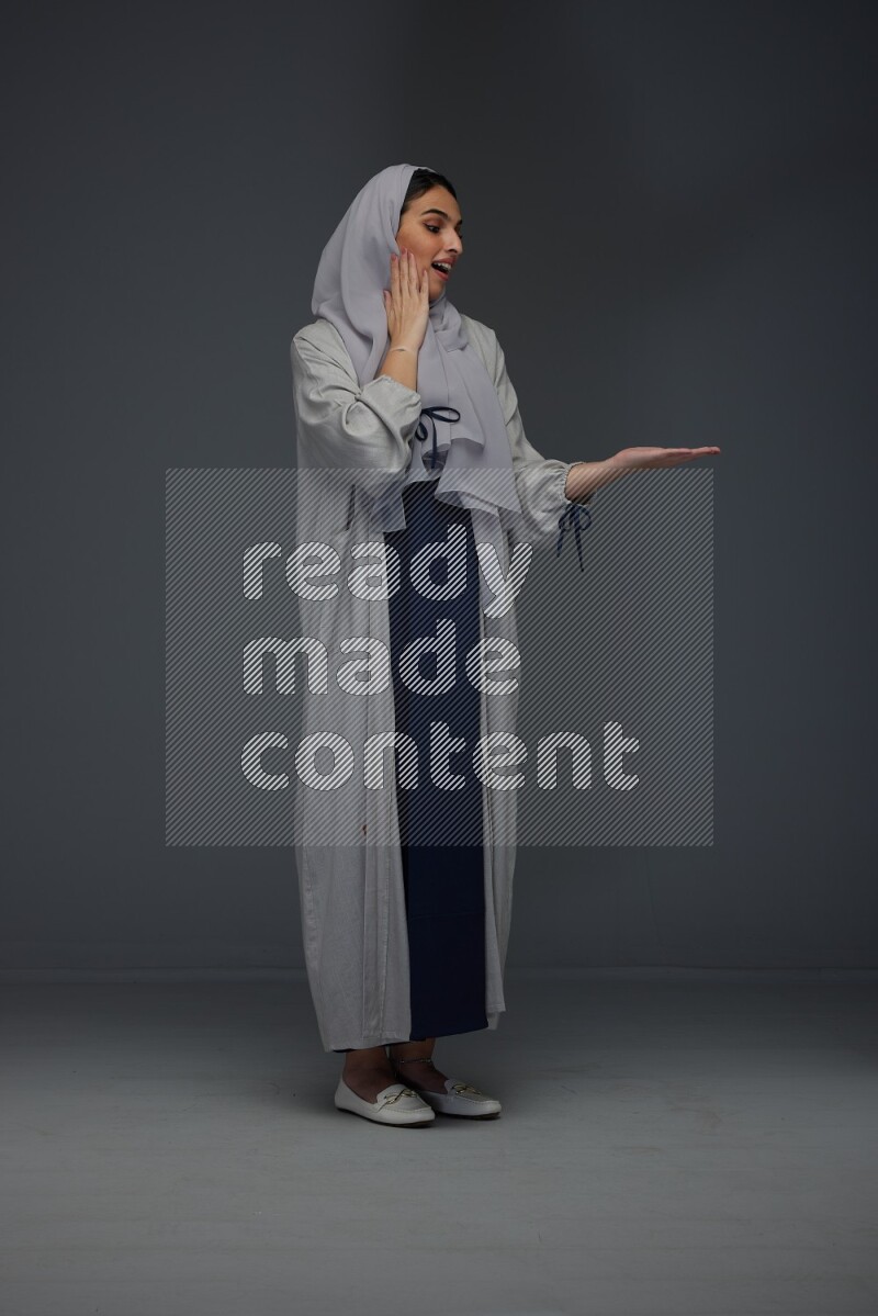 A Saudi woman wearing a light gray Abaya and head scarf standing and pointing in different directions eye level on a grey background