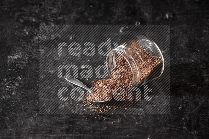 A glass jar full of flaxseeds flipped and seeds spread out with a metal spoon full of the seeds on a textured black flooring