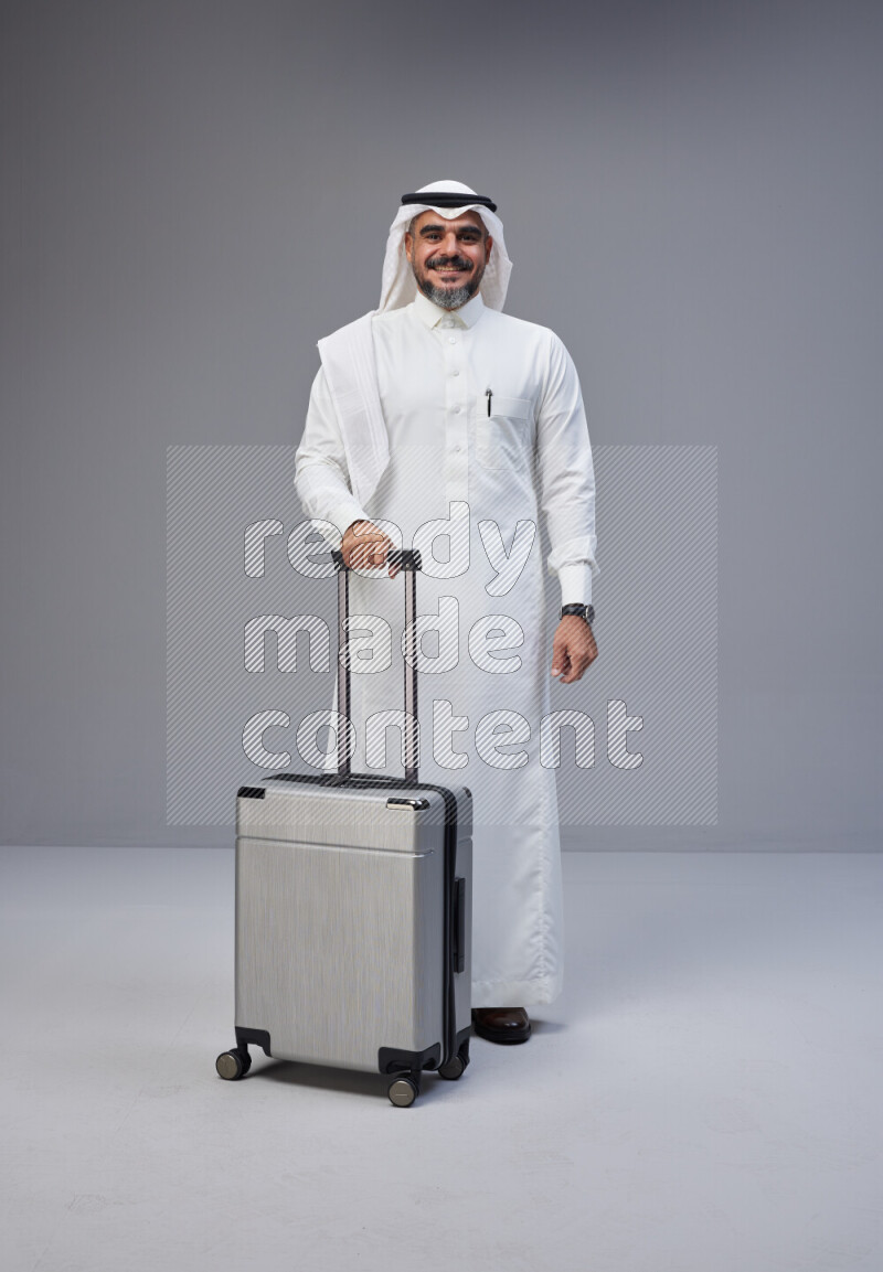 Saudi man wearing Thob and white Shomag standing holding Travel bag on Gray background