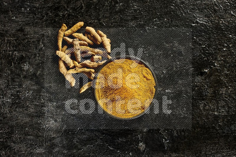 A black pottery bowl full of turmeric powder and dried turmeric whole fingers next of it on textured black flooring