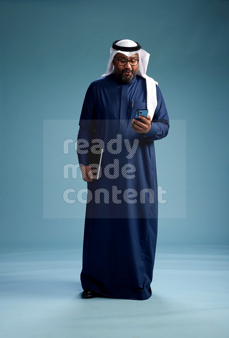 Saudi Man with shimag Standing texting on phone on blue background