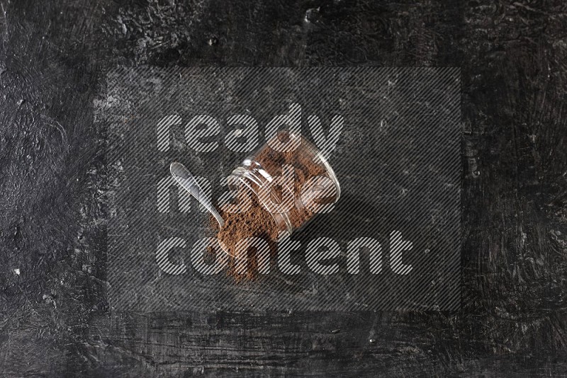 A flipped glass jar and metal spoon full of cloves powder with cloves spread on a textured black flooring