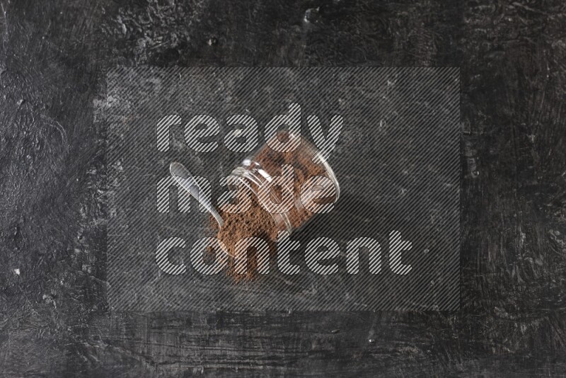 وعاء زجاجي مقلوب وملعقة معدنية ممتلئان ببودرة القرنفل مع حبوب القرنفل المتناثرة على أرضية سوداء