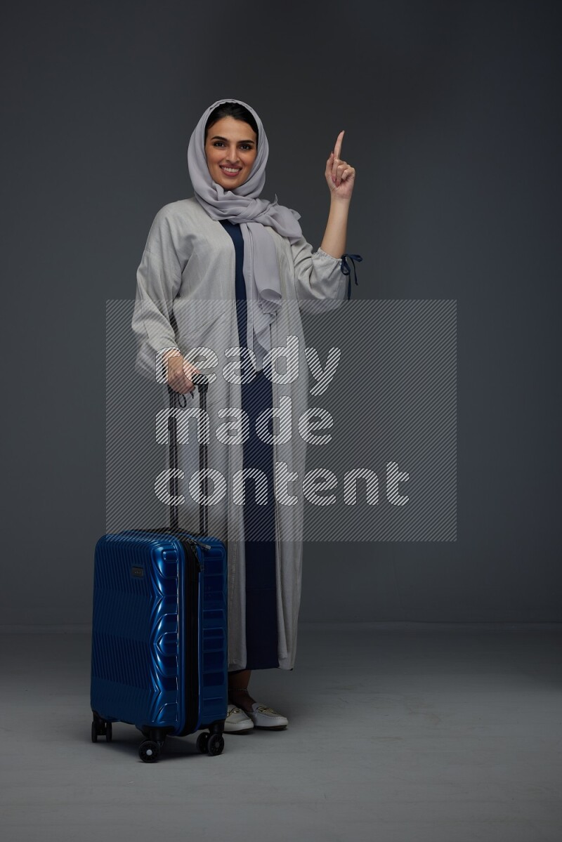 A Saudi woman wearing a light gray Abaya and head scarf standing while holding blue luggage eye level on a grey background