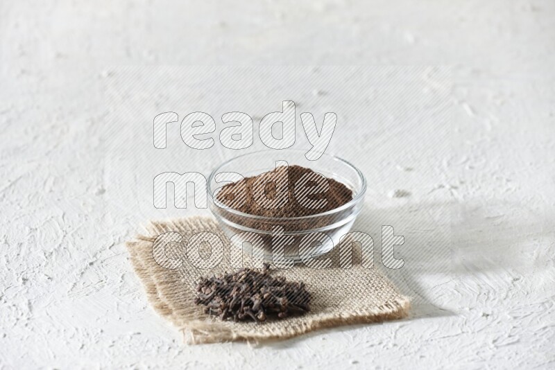 A glass bowl full of cloves powder and cloves grains on a burlap piece on a white flooring