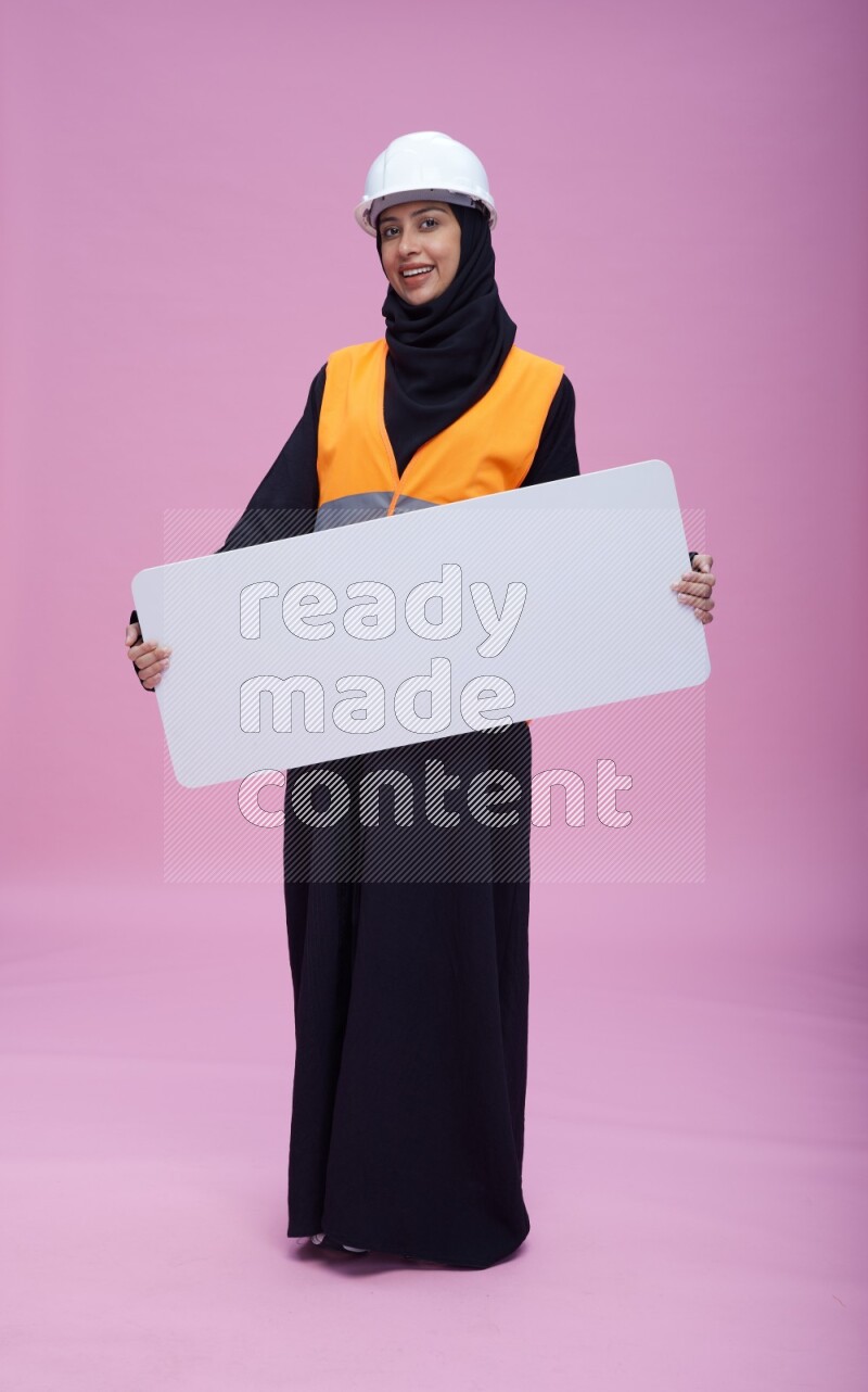 Saudi woman wearing Abaya with engineer vest and helmet standing holding board on pink background