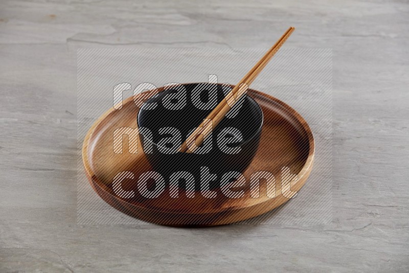 black ceramic round bowl on top of brown wood round plate and wood chopsticks, on grey textured countertop