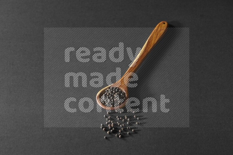 A wooden ladle full of black pepper beads on a black flooring