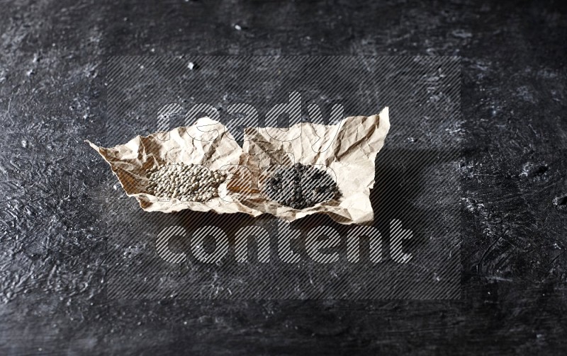 2 Crumpled pieces of paper full of black and white pepper beads on a textured black flooring