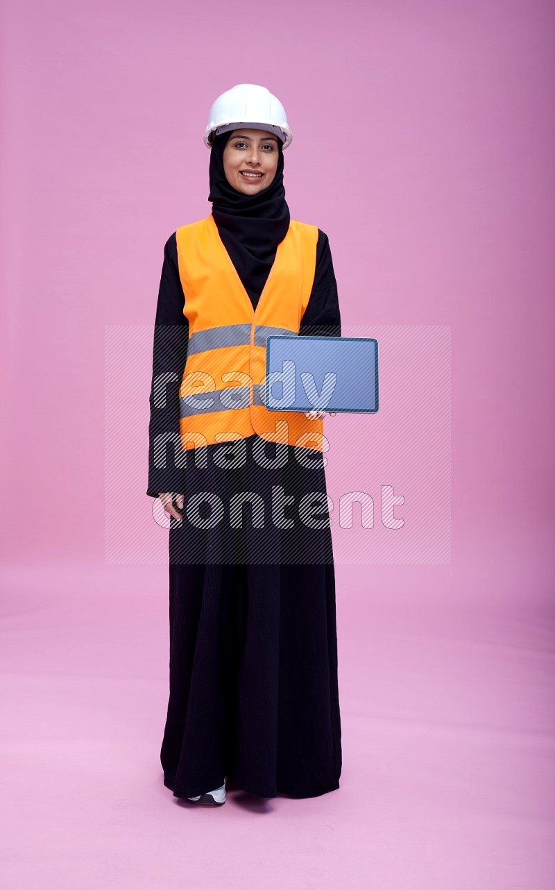 Saudi woman wearing Abaya with engineer vest and helmet standing showing tablet to camera on pink background