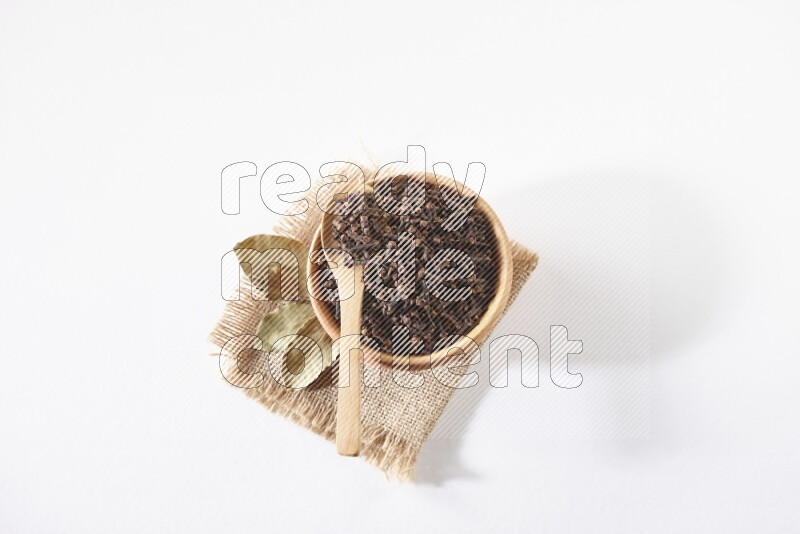 A wooden bowl and a wooden spoon full of cloves on a piece of burlap on a white flooring