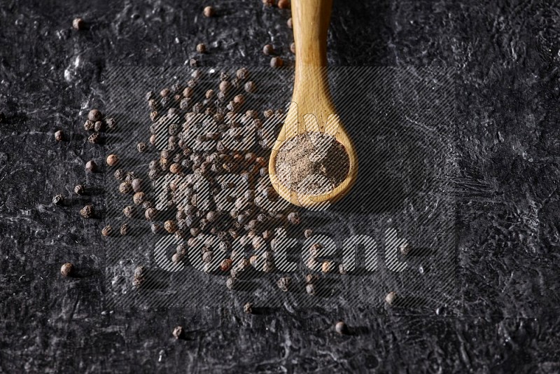A wooden spoon full of black pepper powder and black pepper beads spread on a textured black flooring