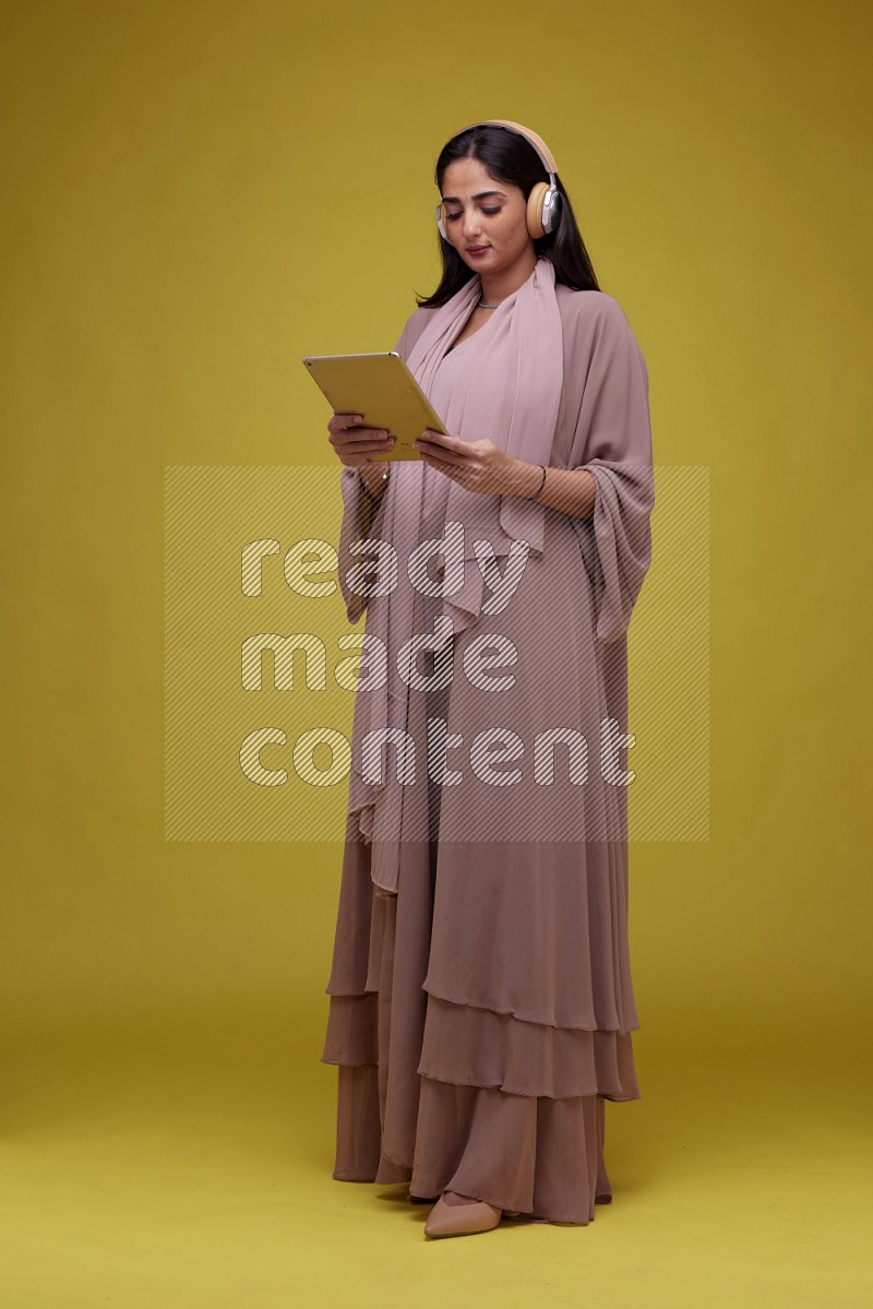 A woman Holding an Ipad with Headphone on a Yellow Background wearing Brown Abaya