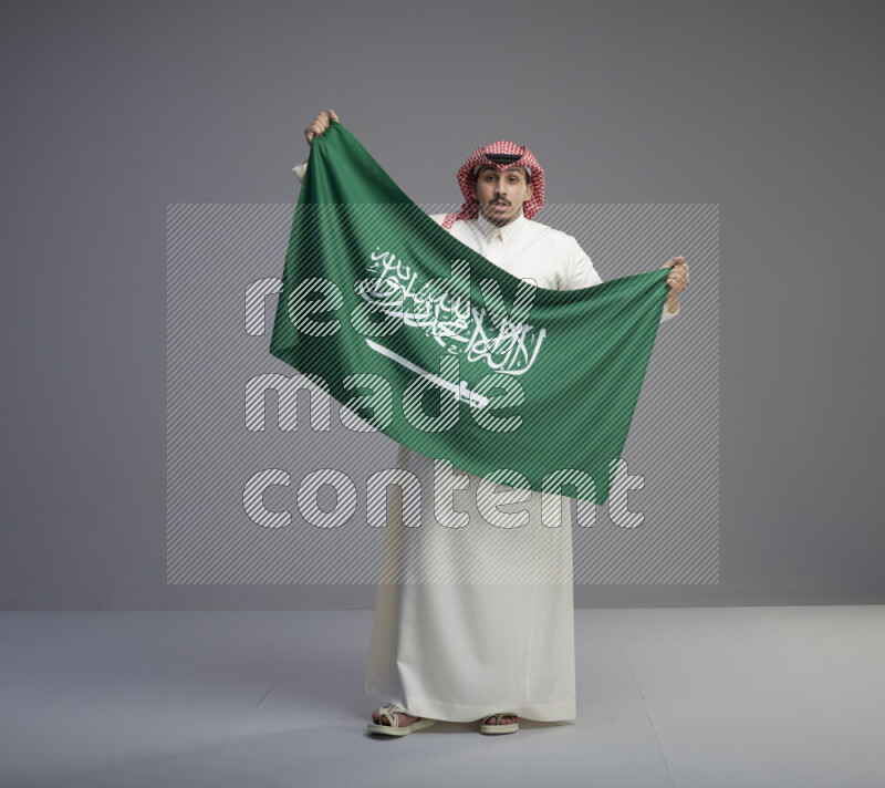 A saudi man standing wearing thob and red shomag holding big saudi flag on gray background