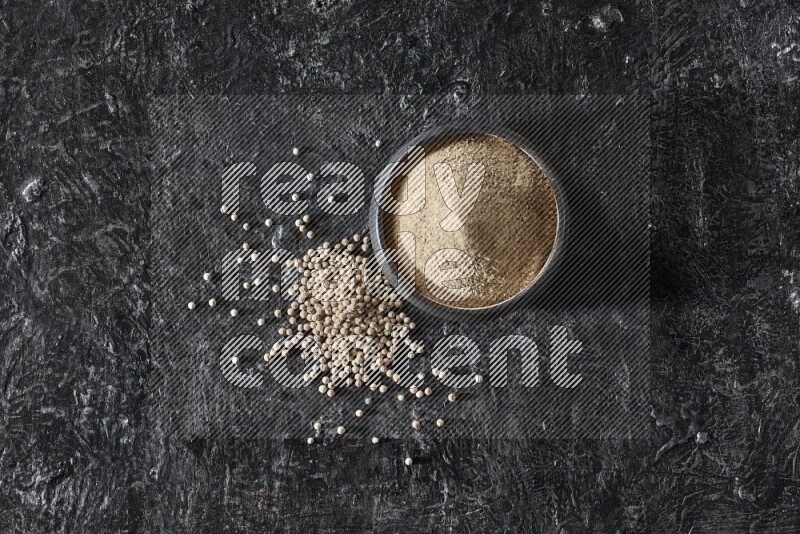 Black pottery bowl full of white pepper powder with white pepper beads on textured black flooring