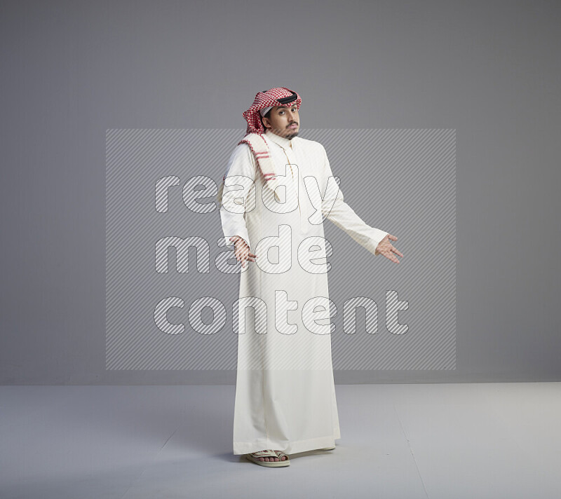 A Saudi man standing wearing thob and red shomag interacting with the camera on gray background