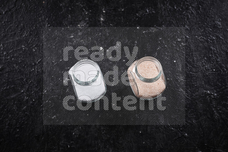 2 وعاء زجاجي احدهمها ممتلئ بملح الهيمالايا الناعم والأخر بملح الطعام على خلفية سوداء