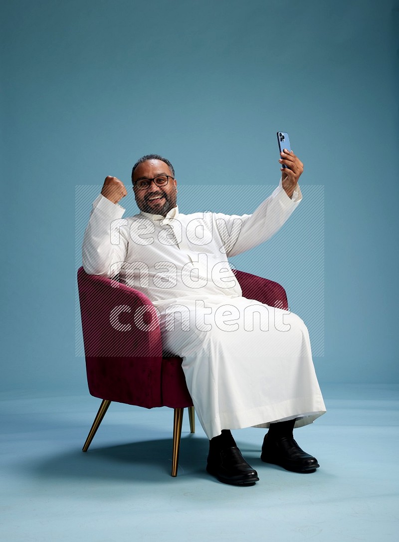 Saudi Man without shimag sitting on chair taking selfie on blue background