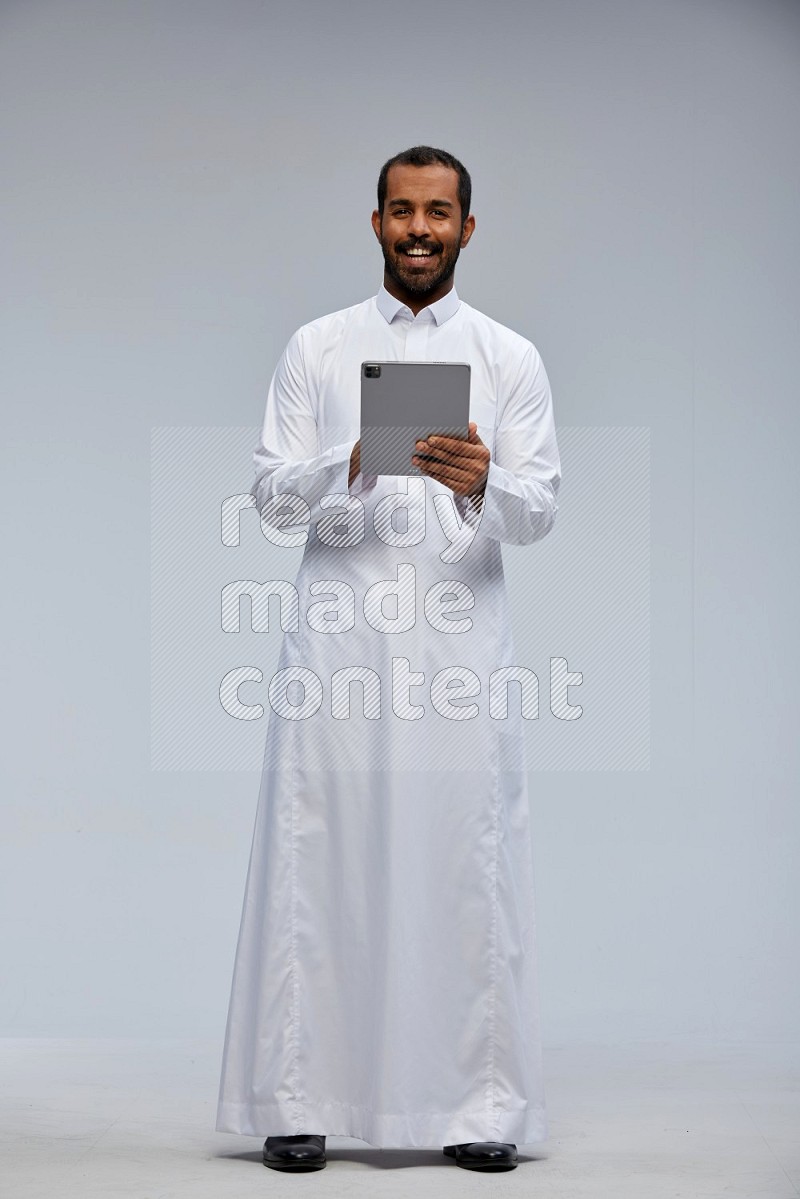 Saudi man wearing Thob standing working on tablet on Gray background