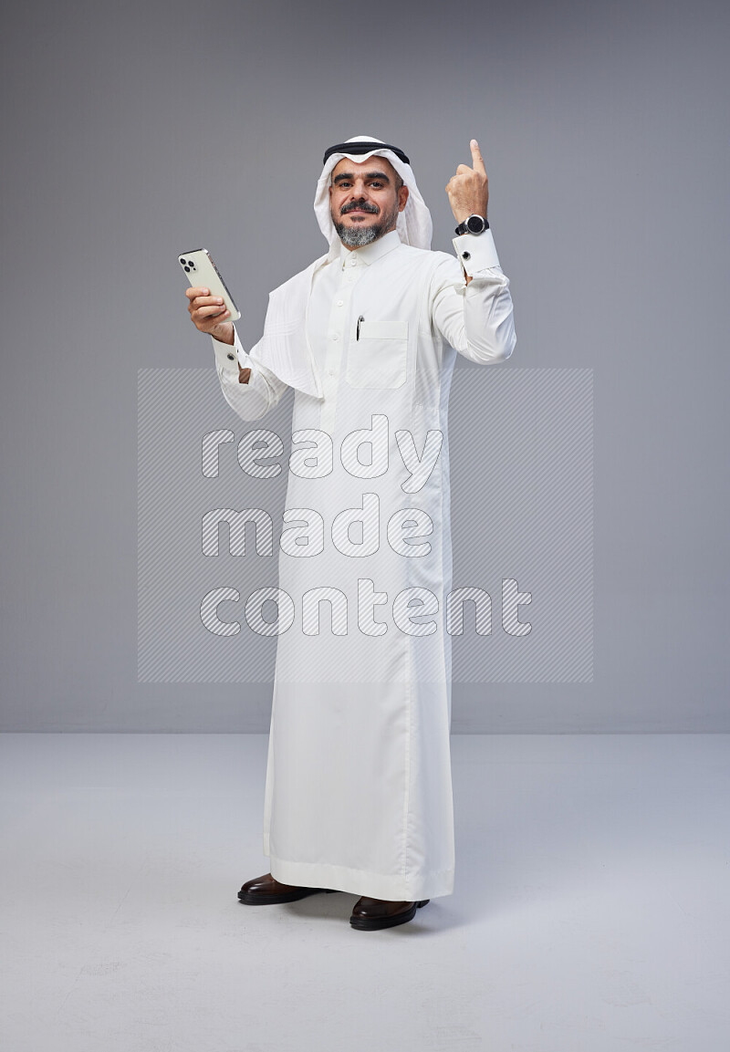 Saudi man Wearing Thob and white Shomag standing texting on phone on Gray background