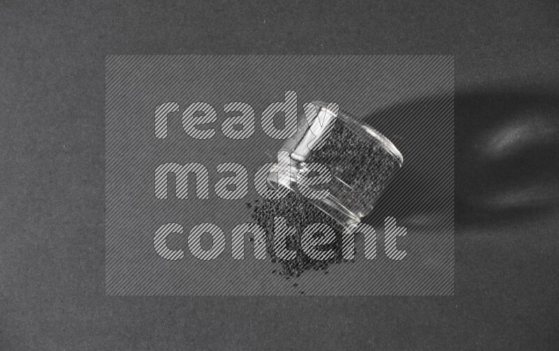 وعاء زجاجي مقلوب ممتلئ ببذور حبة البركة والبذور متناثرة على أرضية سوداء