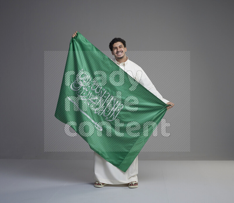 A saudi man standing wearing thob holding big saudi flag on gray background