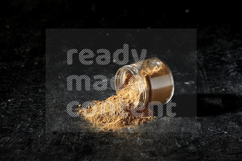 A flipped glass jar full of allspice powder and powder spilled out of it on a textured black flooring