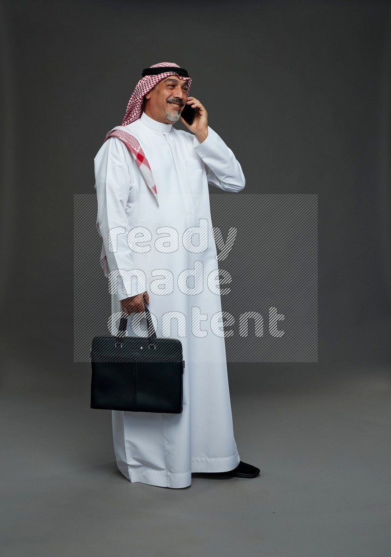 Saudi man with shomag Standing holding bag talking on phone on gray background