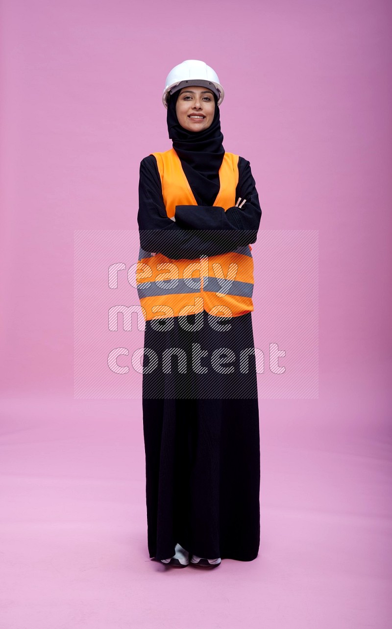 Saudi woman wearing Abaya with engineer vest and helmet standing with crossed arms on pink background