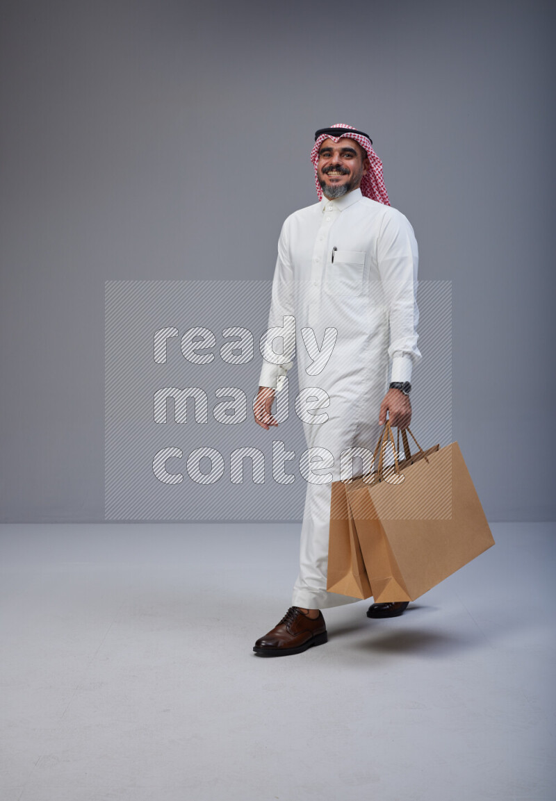 Saudi man Wearing Thob and red Shomag standing holding shopping bag on Gray background