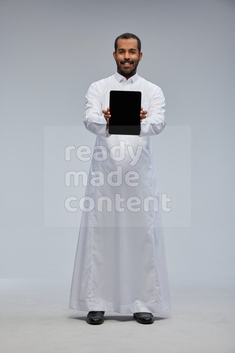 Saudi man wearing Thob standing showing tablet to camera on Gray background