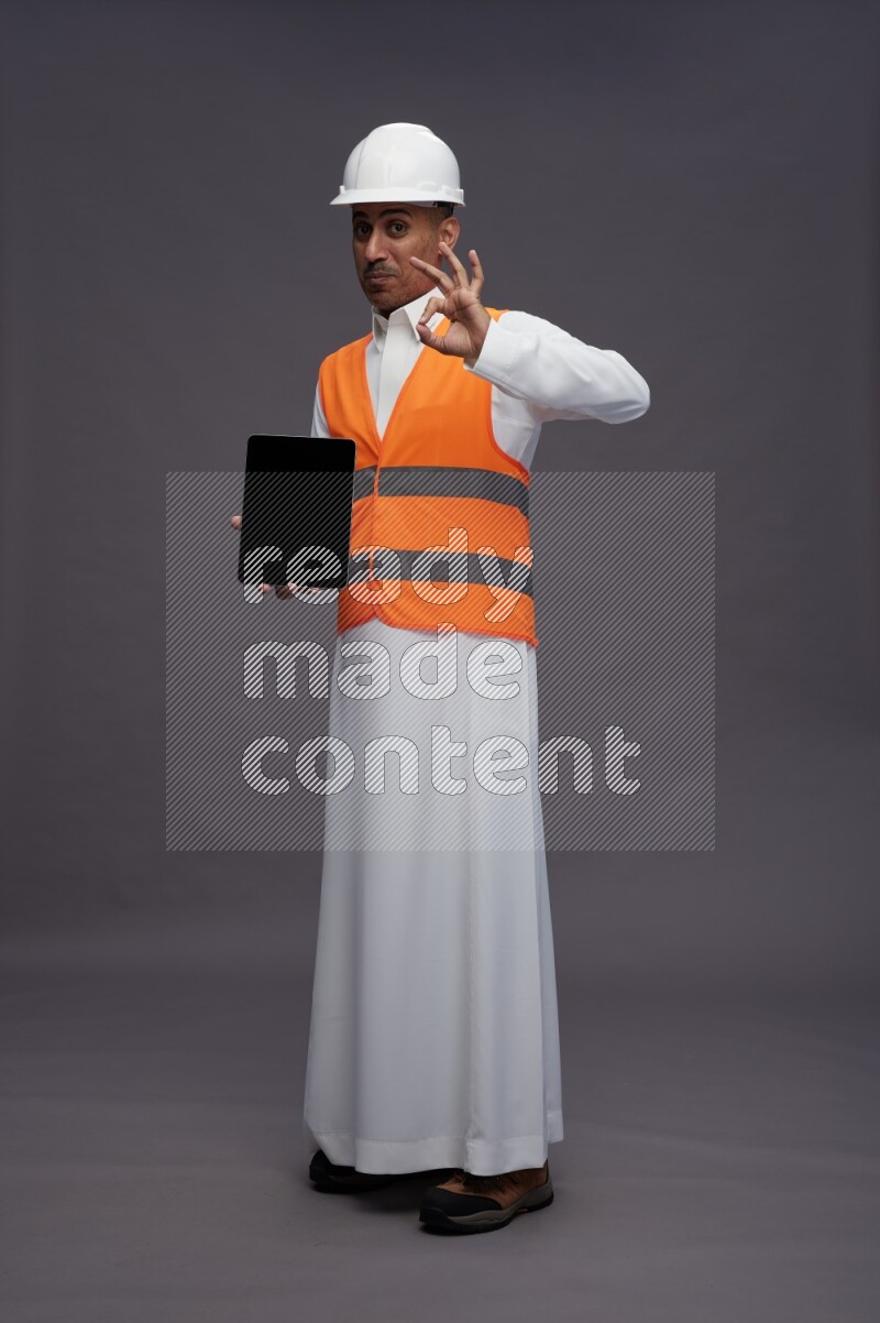 Saudi man wearing thob with engineer vest standing showing tablet to camera on gray background