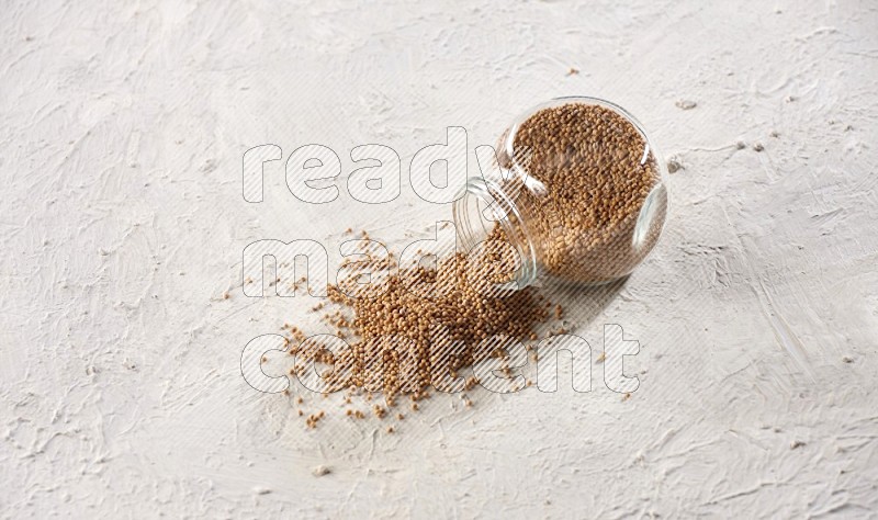 A glass spice jar full of mustard seeds and jar is flipped with fallen seeds on a textured white flooring