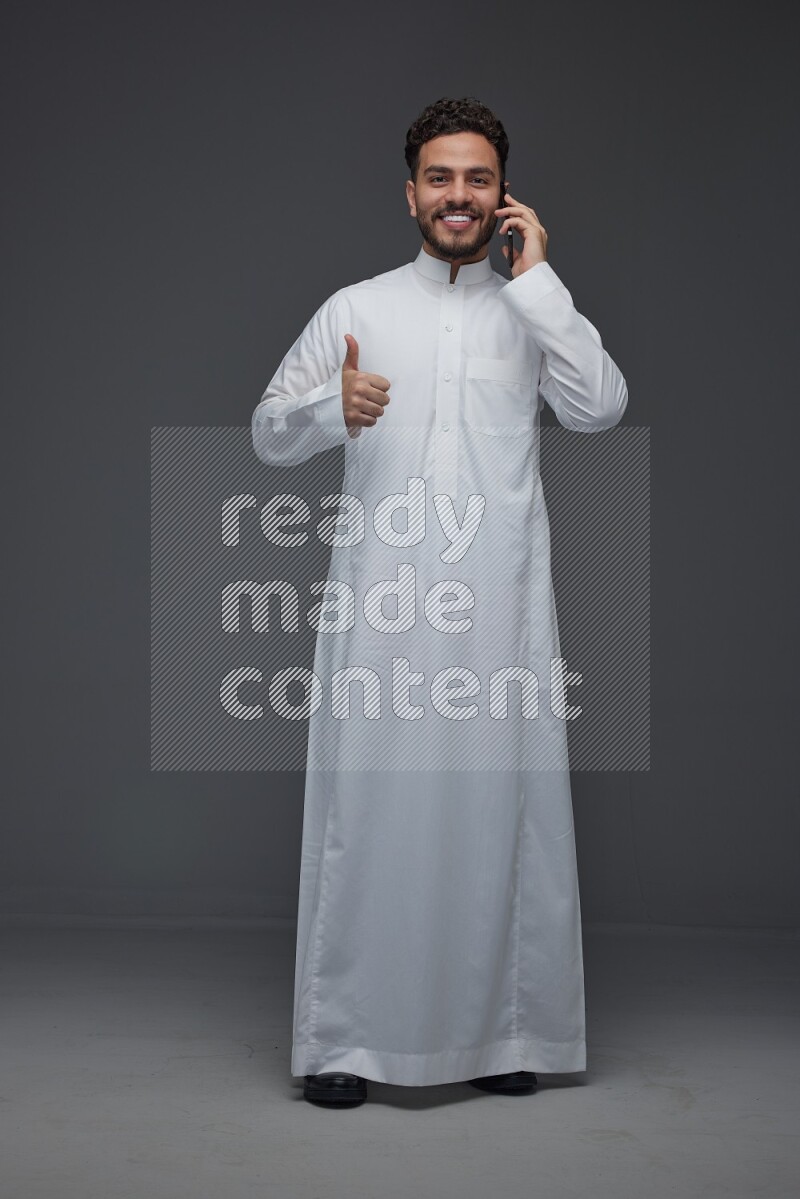 A Saudi man wearing Thobe and talking in the phone while standing and making different hand gestures eye level on a gray background
