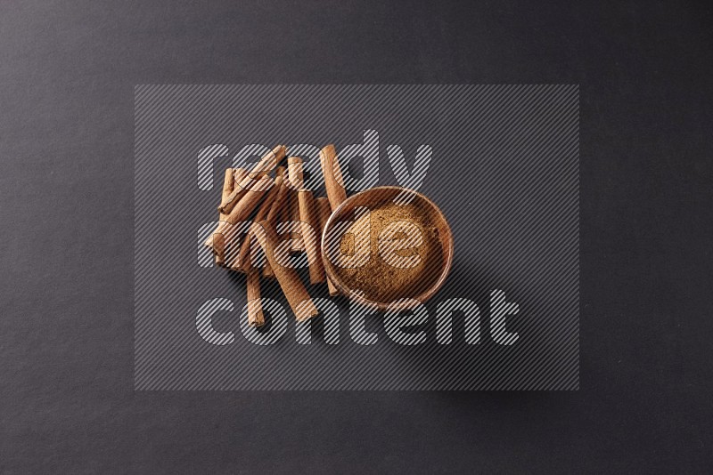 Cinnamon sticks stacked beside a wooden bowl full of cinnamon powder on black background