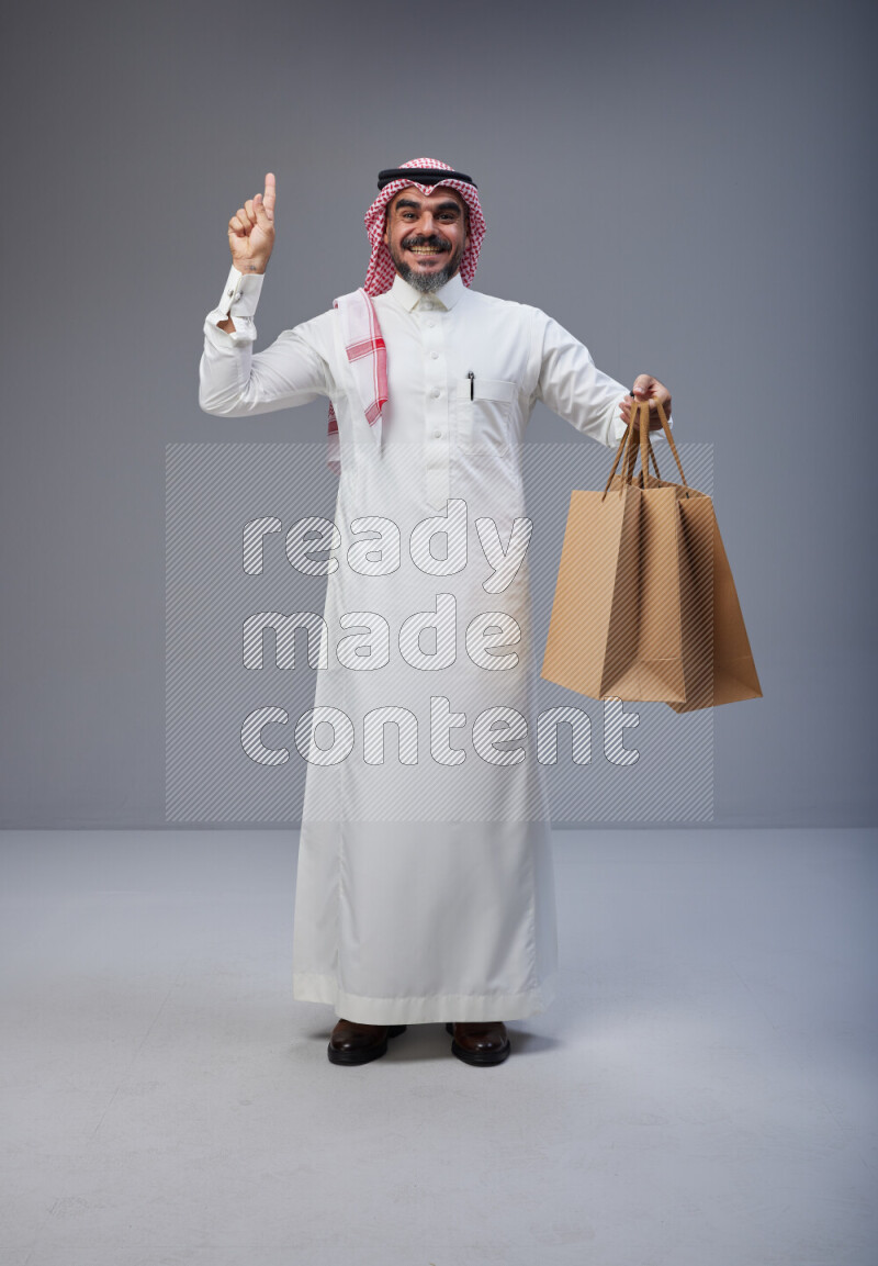 Saudi man Wearing Thob and red Shomag standing holding shopping bag on Gray background
