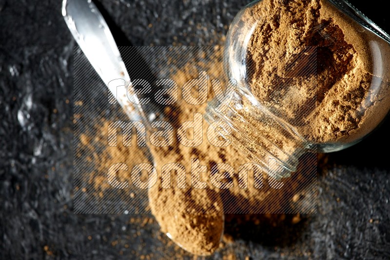 A flipped glass spice jar and a metal spoon full of allspice powder and powder spilled out of it on a textured black flooring