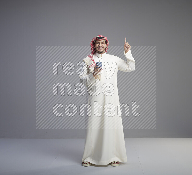 A  Saudi man standing wearing thob and red shomag texting on phone on gray background