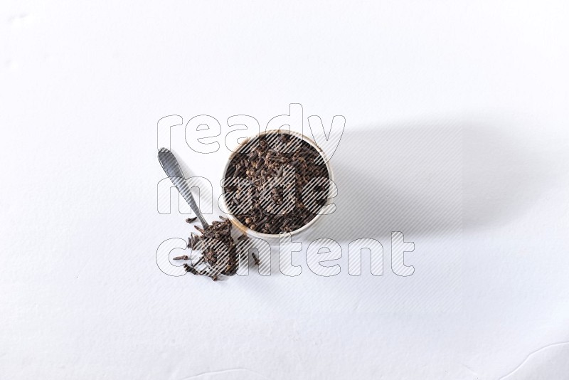 A beige ceramic bowl full of cloves and a metal spoon next to it on a white flooring