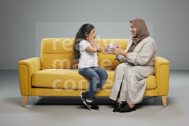 A girl sitting on a yellow sofa and giving a gift to her mother on gray background