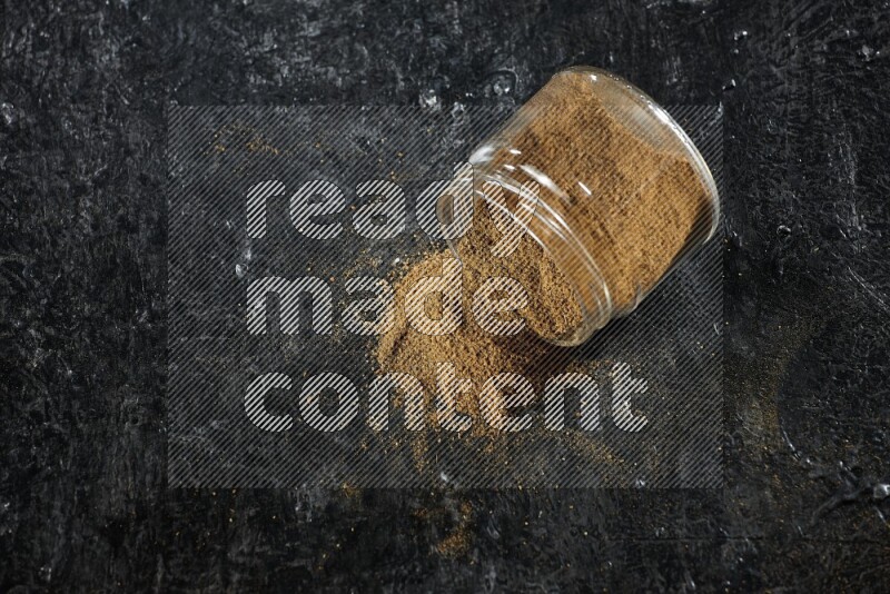 وعاء زجاجي مقلوب ممتلئ ببودرة الكمون والبودرة تناثرت منه على خلفية سوداء