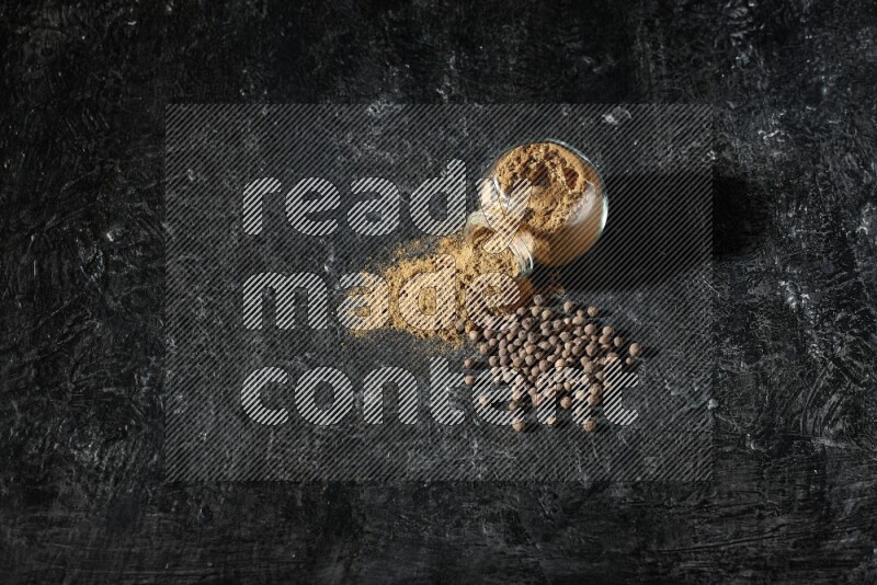 وعاء زجاجي للتوابل مقلوب ممتلئ ببودرة بهار الفلفل الحلو والبودرة تناثرت منه مع الحبوب على خلفية سوداء