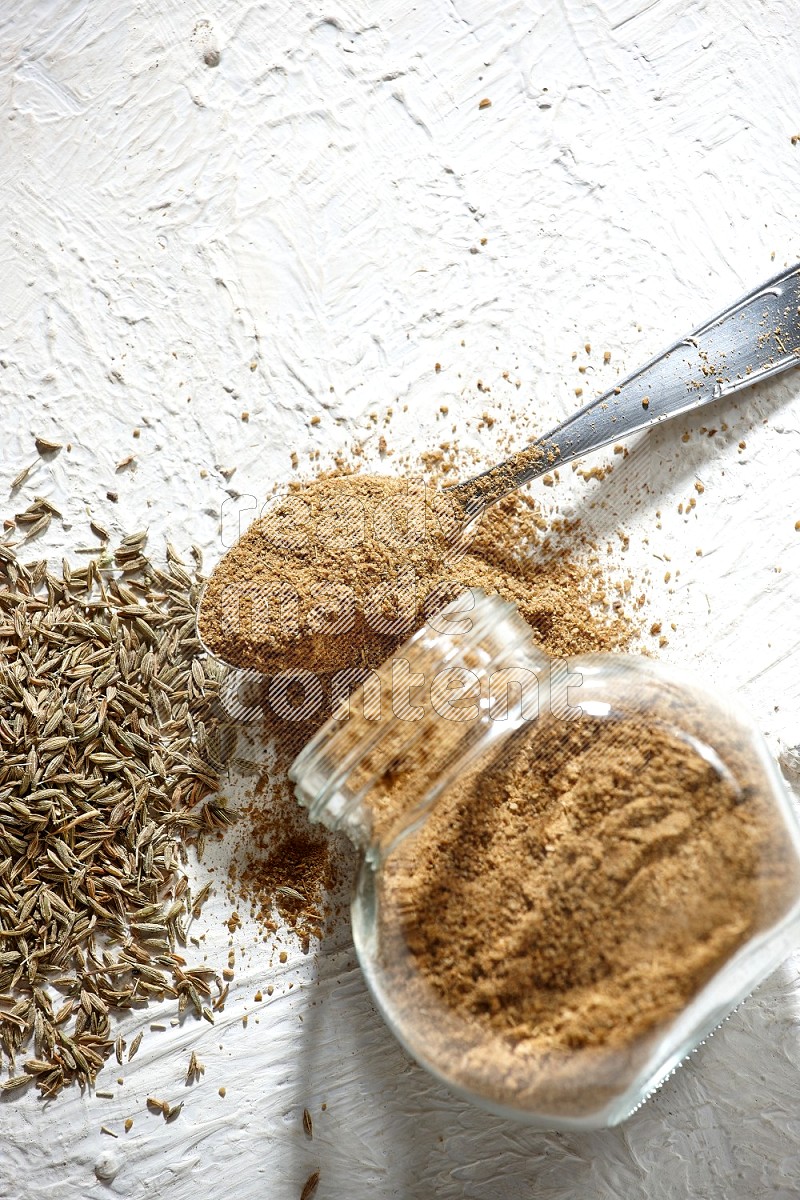 A glass spice jar and metal spoon full of cumin powder and the jar flipped and powder spilled out with cumin seeds on textured white flooring