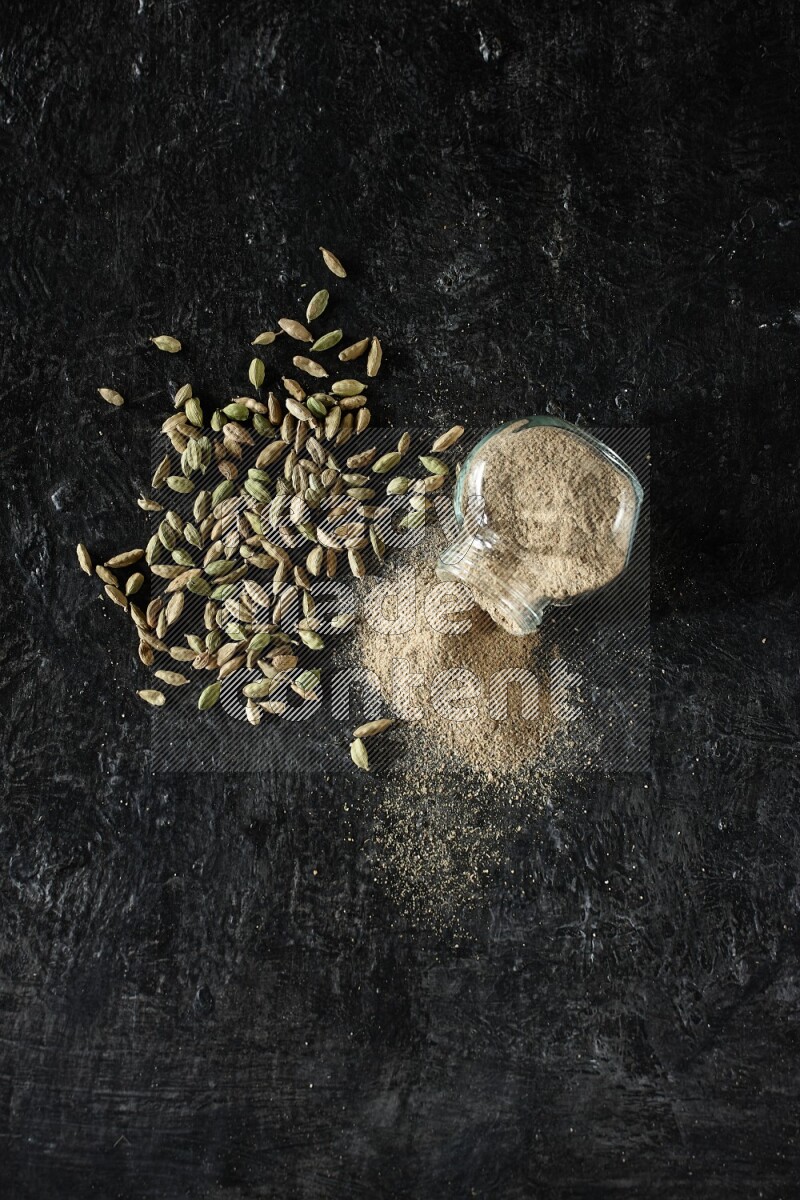 وعاء زجاجي للتوابل مقلوب وممتلئ ببودرة الهيل والبودرة تناثرت منه مع حبوب الهيل المتناثرة على خلفية سوداء