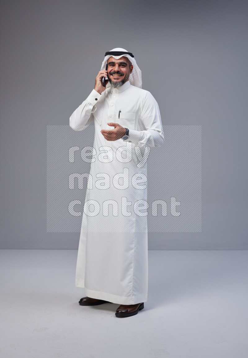 Saudi man Wearing Thob and white Shomag standing talking on phone on Gray background