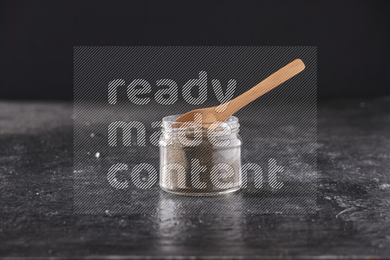 A glass jar full of black pepper powder and a wooden spoon on a textured black flooring