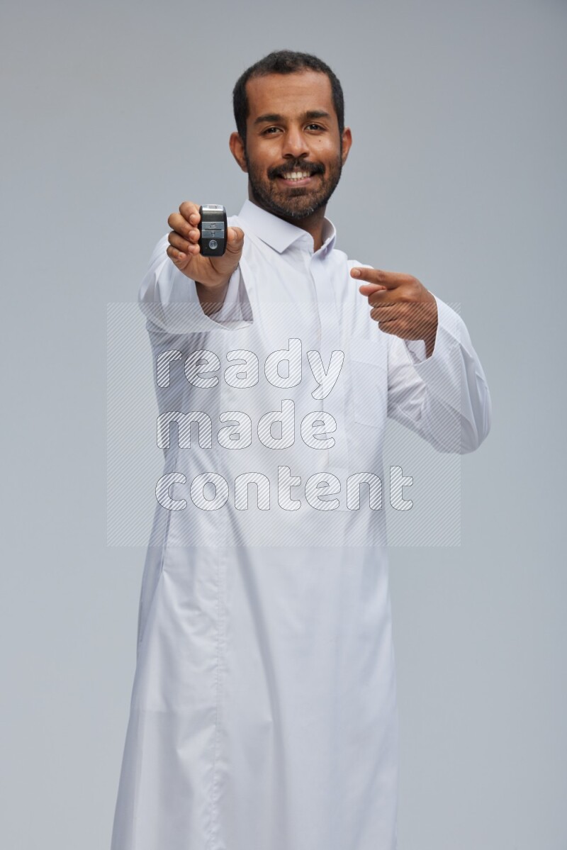 Saudi man wearing thob and shomag standing holding car key on gray background