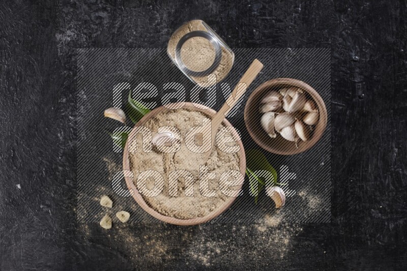A wooden bowl with a spoon in it and glass spice jar all, full of garlic powder and a wooden bowl full of garlic cloves on a black flooring
