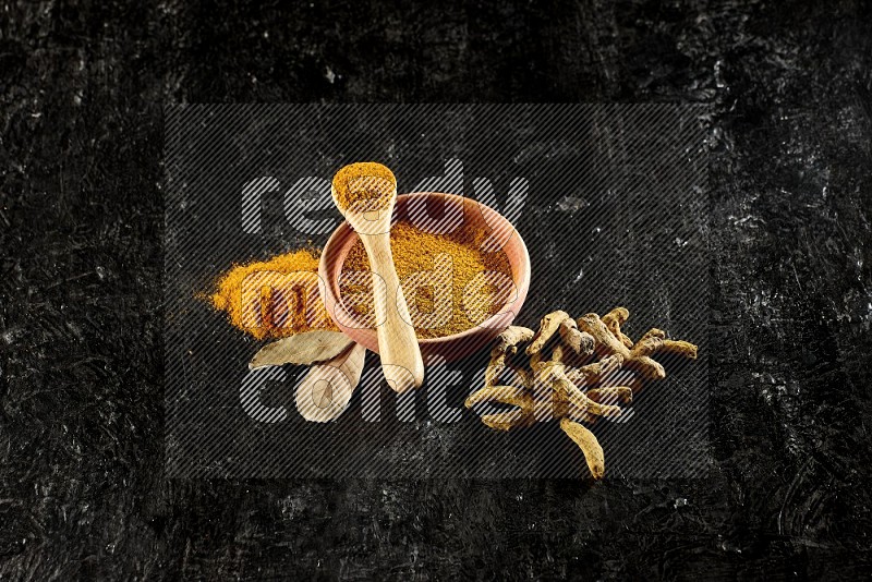 A wooden bowl and a wooden spoon full of turmeric powder with dried turmeric fingers on textured black flooring